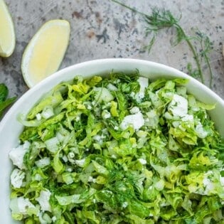 maroulosalata in a white serving bowl next to sprigs of mint and dill, lemon wedges and a bowl of salt with a spoon.