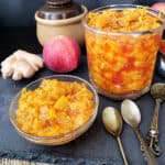 A glass jar and a container filled with apple ginger chutney. There are two apples, piece of ginger and three spoons in the frame.