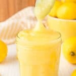 cooked bright yellow lemon curd in a jar with a spoon taking some out with fresh lemons behind the jar.
