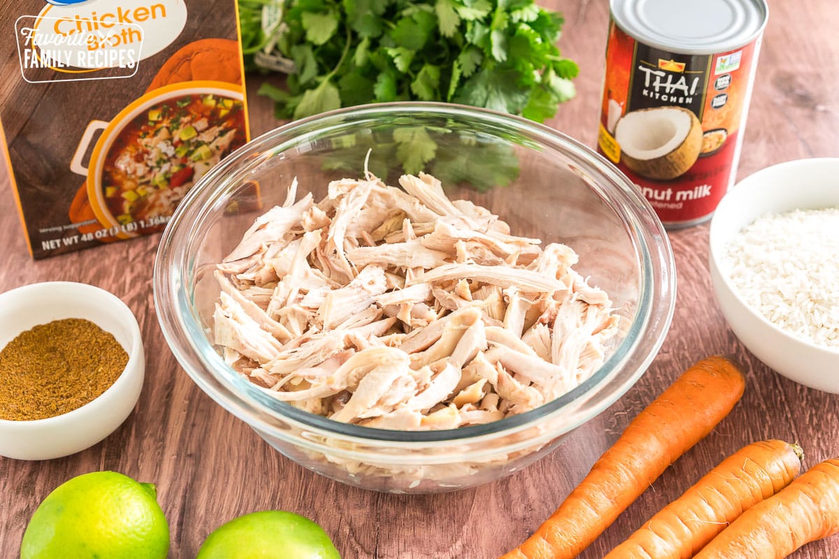 a bowl of shredded chicken surrounded by limes, carrots, rice, chicken broth, coconut milk, and a bowl of thai spices