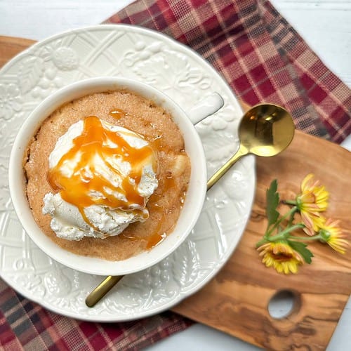 A white mug containing a light brown apple cake topped with a dollop of ice cream and drizzled with caramel sauce. The bowl is placed on a white plate with embossed designs, accompanied by a gold spoon on a wooden board with a red plaid cloth and yellow flowers.