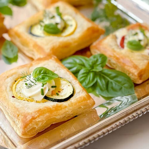 Horizontal closeup view of puff pastry appetizer served on a silver tray with fresh vegetables and herbs.